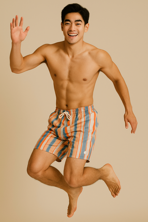 A shirtless man jumping playfully while wearing multicolour striped swim shorts, against a beige studio backdrop.