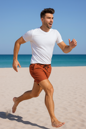 A man running barefoot on the beach wearing terracotta swim shorts and a white t-shirt, with the sea in the background.