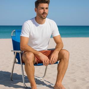 A man sitting on a beach chair, smiling while wearing terracotta swim shorts and a white t-shirt, with turquoise sea behind him.