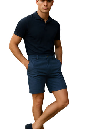 A young man wearing a navy polo shirt, navy tailored shorts, and loafers sits casually on a wooden bench in front of a rustic wooden wall under natural sunlight